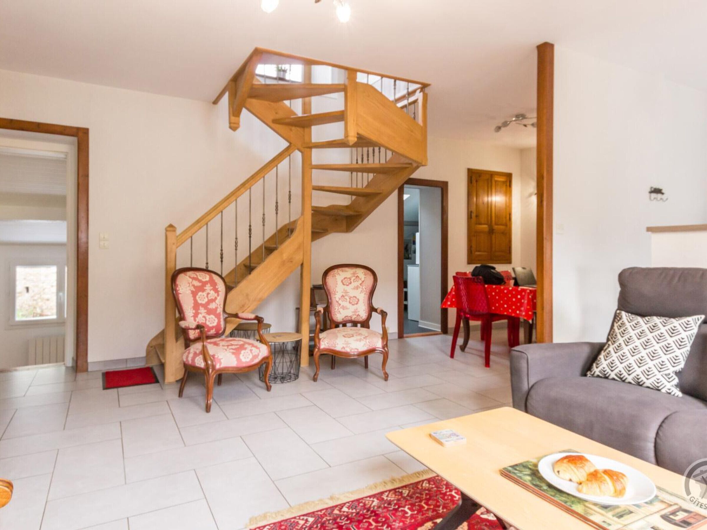 Een woonkamer met een houten trap, twee gestreepte fauteuils, een rode eettafel en een grijze bank.