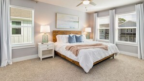 A bedroom with a wooden bed, two bedside tables with lamps, a ceiling fan, and large windows with blinds.