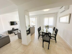 A modern living room with a sofa, a TV, and a dining area with a glass table and chairs.