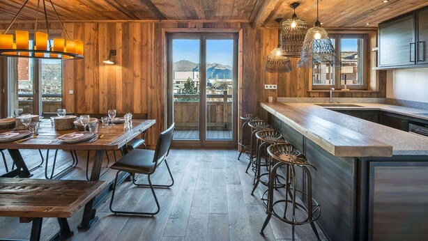 A rustic interior with a wooden ceiling, a dining area with a long table and bar stools, and a kitchen with a large island.