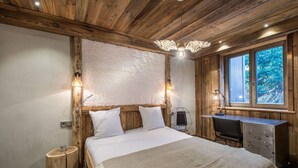 A bedroom with a wooden ceiling, a bed with white bedding, a desk with a chair, and a window with a view of trees.