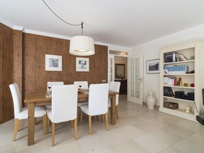Een eetruimte met een houten tafel, witte stoelen, een hangende lamp en een boekenkast met boeken en decoratieve objecten.