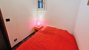 Quarto pequeno com uma cama de solteiro, mesa de cabeceira com abajur e uma janela.