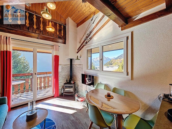 A cozy room with a wooden ceiling, a fireplace, a dining table with chairs, and large windows offering mountain views.