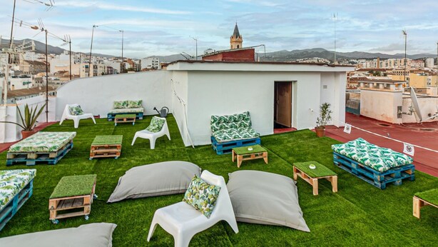 Una terraza en la azotea con césped artificial verde, sillas de plástico blanco y cojines de colores azules y verdes.