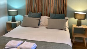 Una cama bien hecha con dos almohadas, una mesita de noche con una lámpara y una toalla doblada en el suelo.
