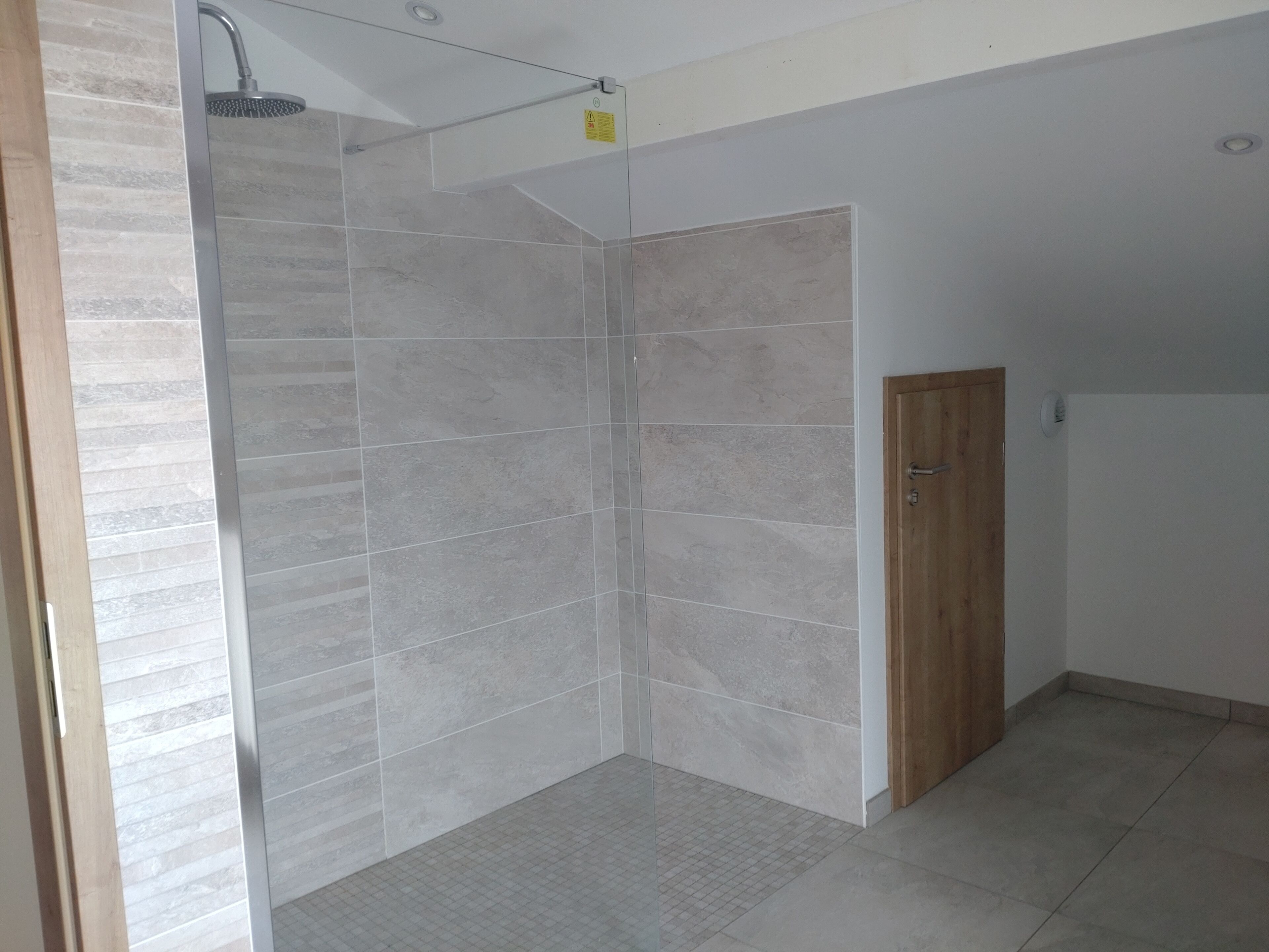 Ein Duschbereich mit gefliesten Wänden und Boden, einer Holztür und einem sichtbaren Deckenventil.