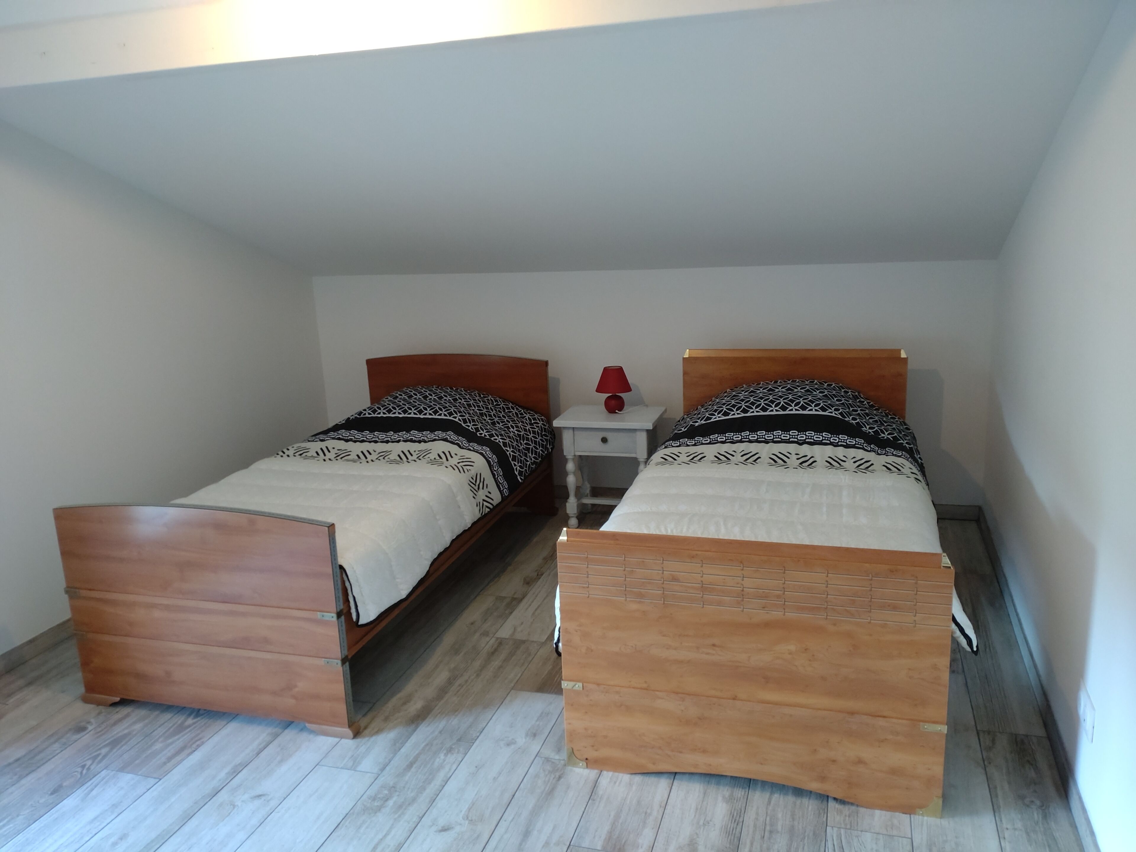Une petite chambre à coucher composée d’une seule pièce, avec deux lits en bois, une table de chevet avec une lampe et un mur blanc.