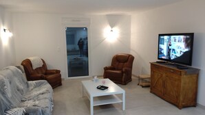 A living room with a brown leather sofa, a white coffee table, a brown leather armchair, a wooden cabinet with a television, and a sliding glass door leading to a balcony.