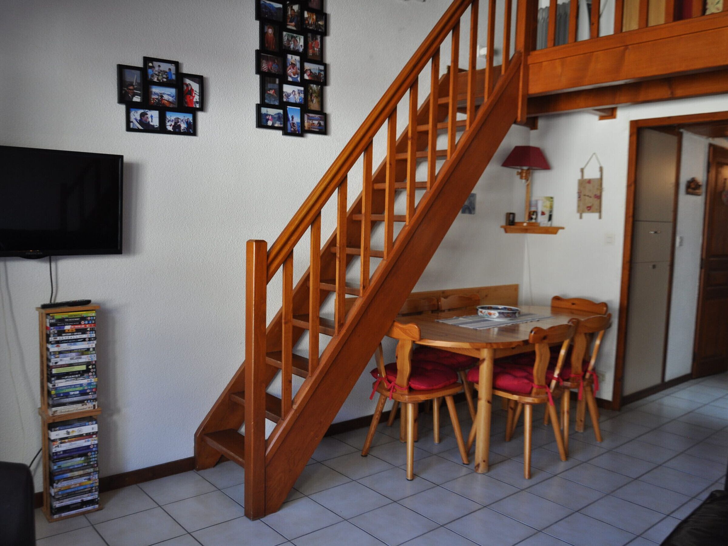 Un escalier menant à un niveau supérieur, une table à manger avec des chaises, un téléviseur et une étagère contenant des DVD.