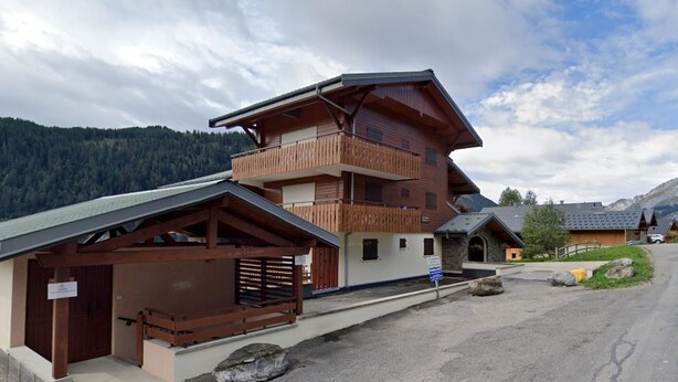 Un edificio de madera de dos pisos con un balcón, una entrada cubierta y montañas al fondo.
