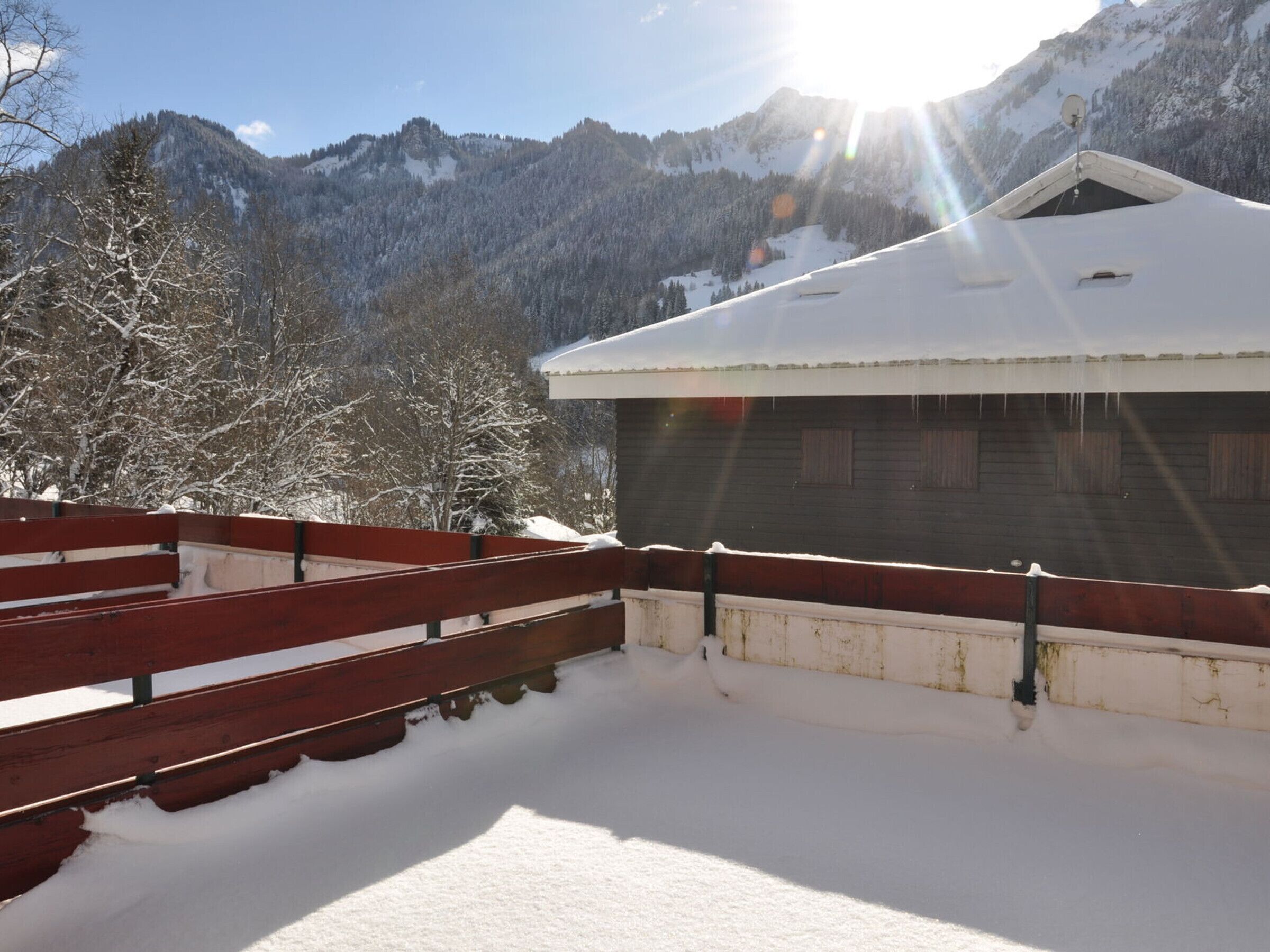 A wooden cabin with a snow-covered roof, surrounded by a red wooden fence and snow-covered trees.