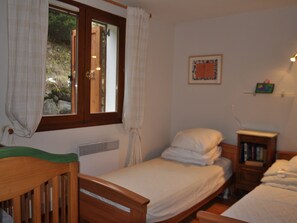 A bedroom with two single beds, a wooden chair, a small bedside table with a lamp, and a window with curtains.