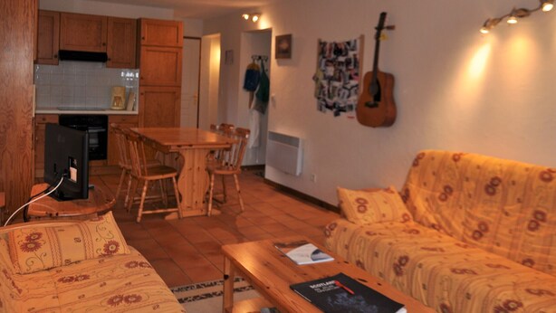 Un salon chaleureux avec un canapé à motifs, une table basse en bois et un coin repas comprenant une table et des chaises en bois. Une guitare est accrochée au mur, et une housse de guitare est posée contre le mur.