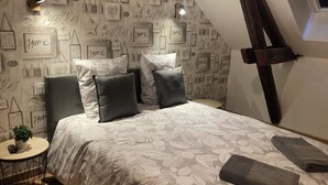 A bedroom with a bed, pillows, a small table with a plant, and a wall with various framed items and text. - La fine bulle! (Épernay)