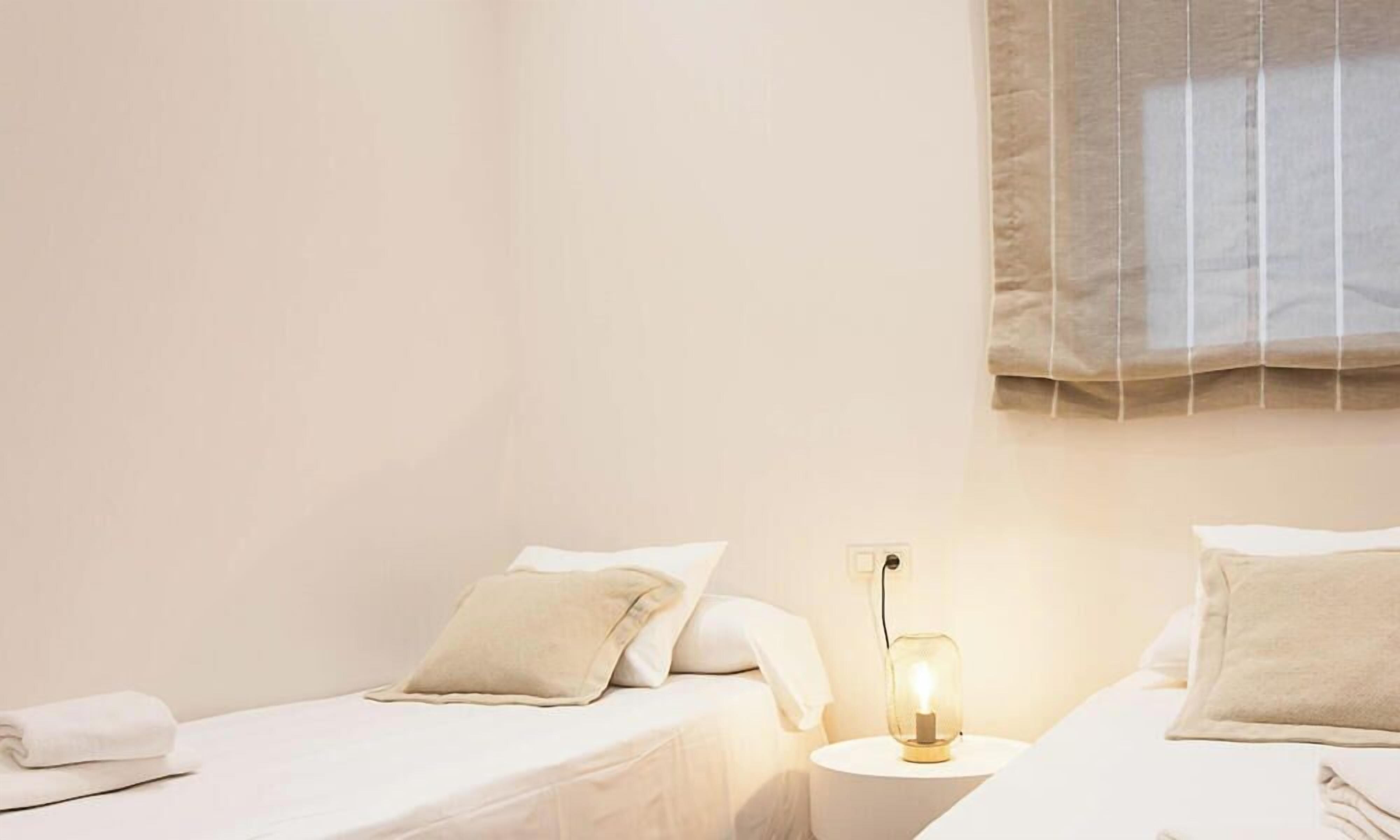 A hotel room with two beds, a bedside table with a lamp, and a window with blinds.