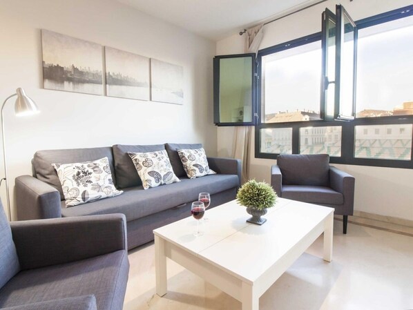 A modern living room with a grey sofa, two armchairs, a white coffee table, and a potted plant. There are three framed artworks on the wall and a large window with a view of buildings.