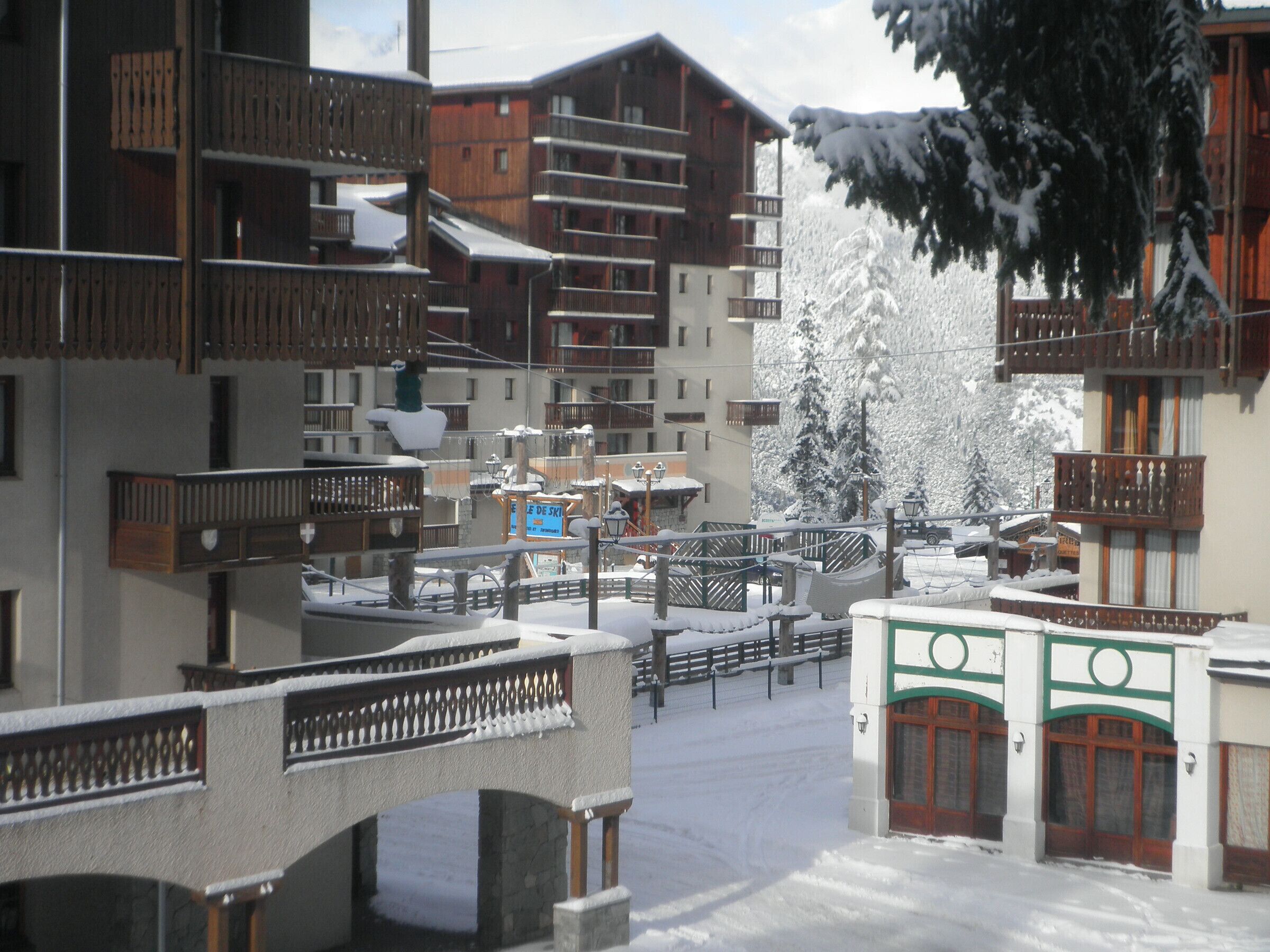 A snow-covered ski resort with multi-story buildings, wooden balconies, and a bridge.