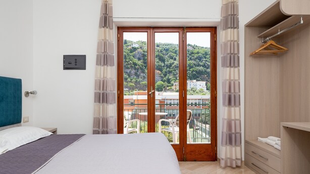 A bedroom with a bed, a wooden wardrobe, a balcony with a view of buildings and greenery. - Parsano suite (Sorrento)
