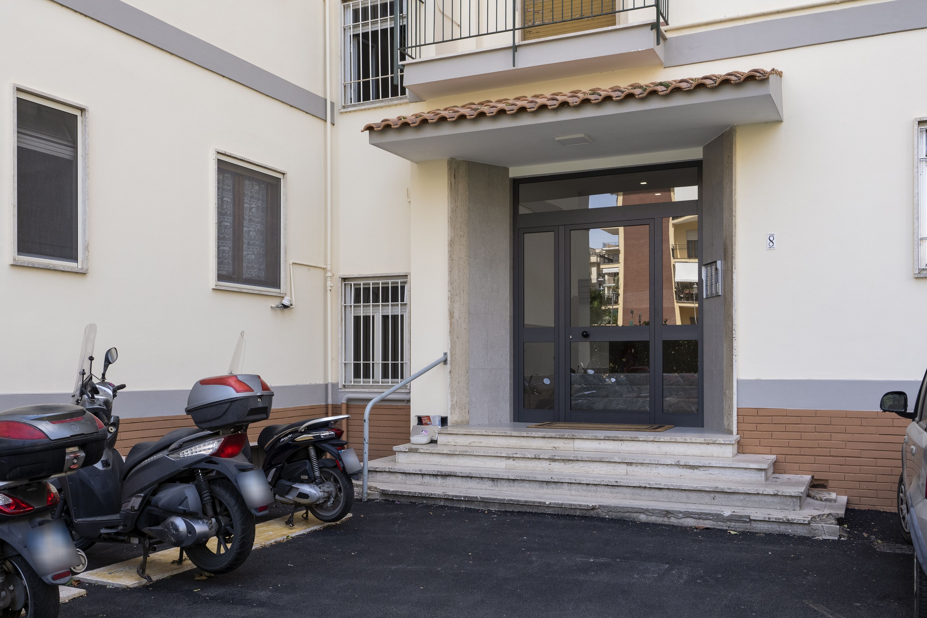 A building entrance with a tiled roof, a set of steps leading to a glass door, and two parked scooters.
