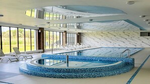 A modern indoor swimming pool with a blue mosaic design, surrounded by lounge chairs and large windows offering a view of the outdoors.