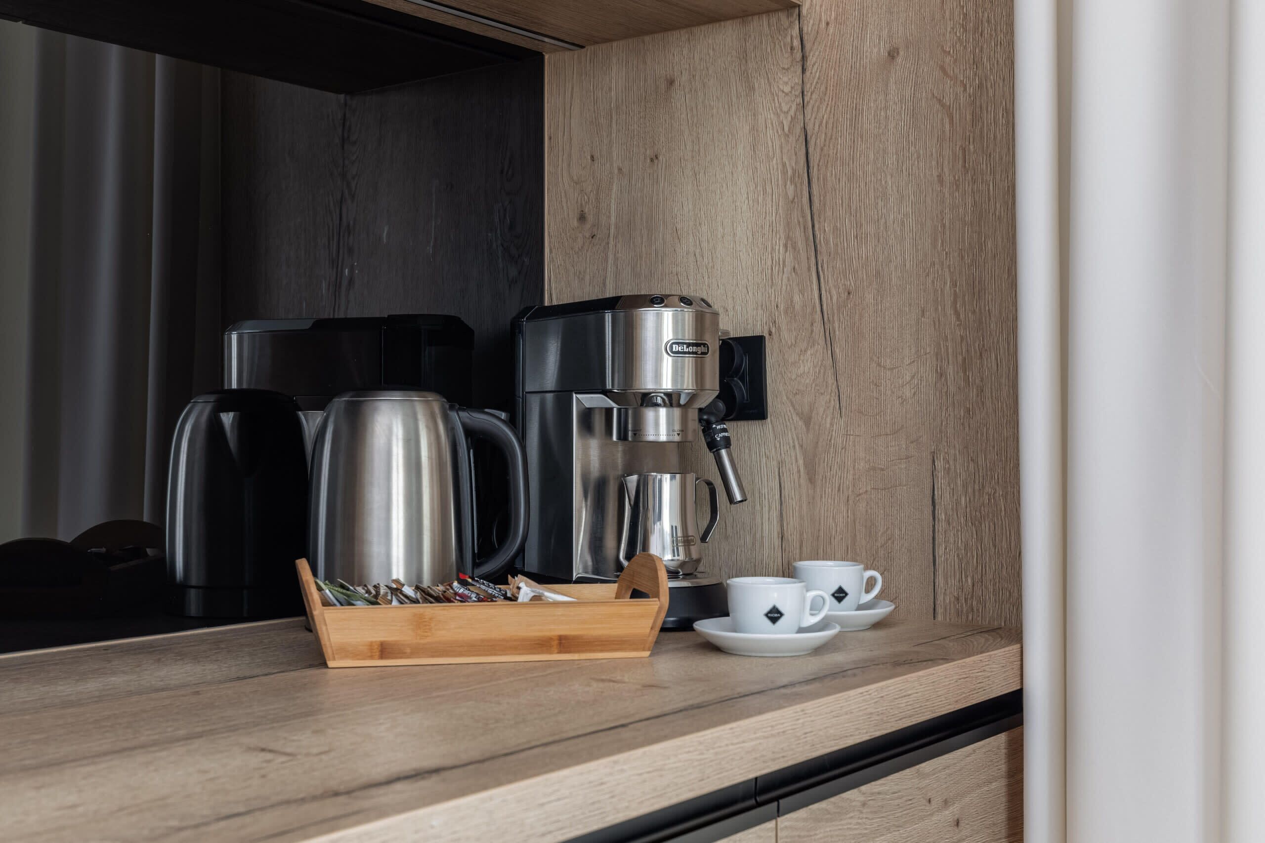 A coffee maker, two cups, and a tray of coffee beans on a wooden countertop.