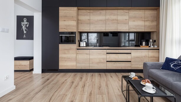 A modern kitchen with wooden cabinets, a black countertop, and a grey sofa with a blue pillow.