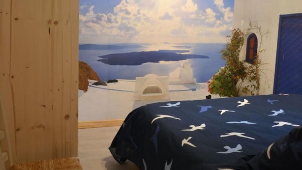 A room with a bed, a view of a coastal landscape with white buildings and the sea, and a blue door.