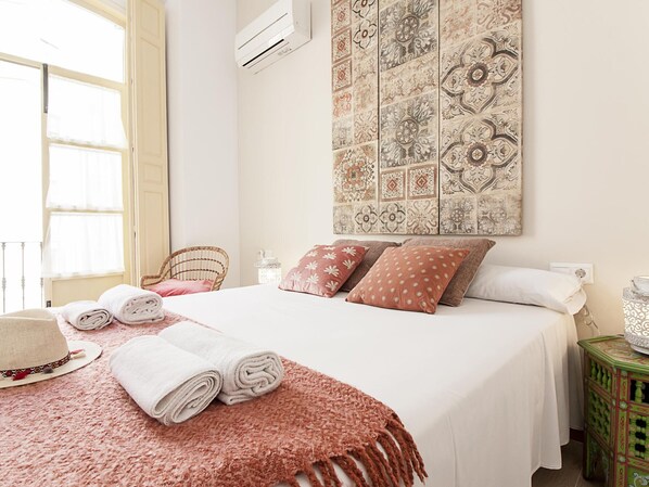 A neatly made bed with rolled towels and a hat, a wicker chair, a decorative wall panel, and a small table with a candle.