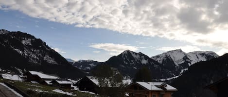 A mountain village with wooden houses, a wooden bridge, and snow-covered mountains.