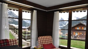 A cozy room with a checkered armchair, a sofa, and a small table with books. Large windows offer a view of mountains and a balcony. - Apartment with terrace, ski locker and covered parking (Châtel)