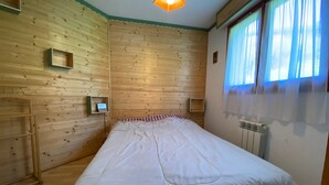 Ein Zimmer mit Holzwänden, einem Bett mit weißer Bettdecke, zwei Nachttischen, einem Fenster mit Vorhängen und einem Heizkörper.