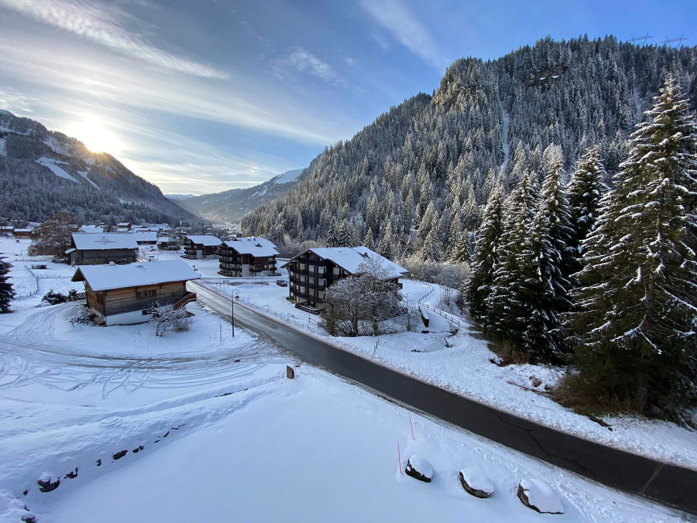 Ein verschneites Dorf mit Holzhäusern, einer kurvenreichen Straße und einem bewaldeten Berg im Hintergrund.