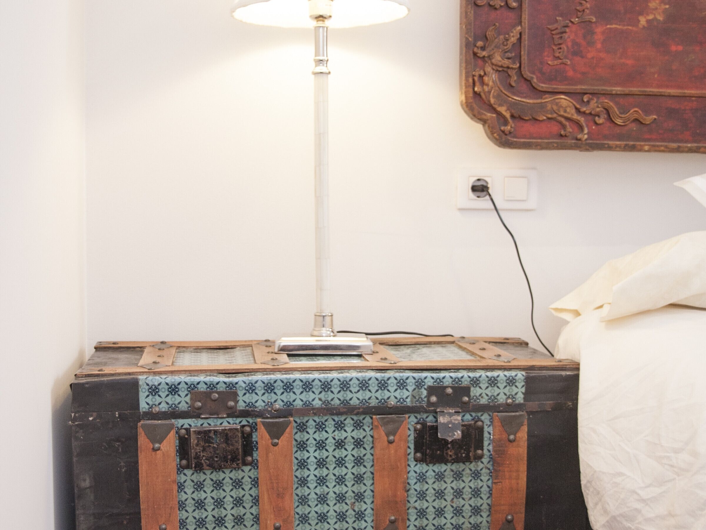 A bed with a white pillow, a lamp on a wooden chest, and a wall-mounted decorative item.