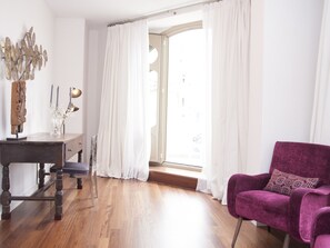 Une pièce avec un plancher en bois, un bureau avec une lampe, une chaise et une fenêtre avec des rideaux blancs.