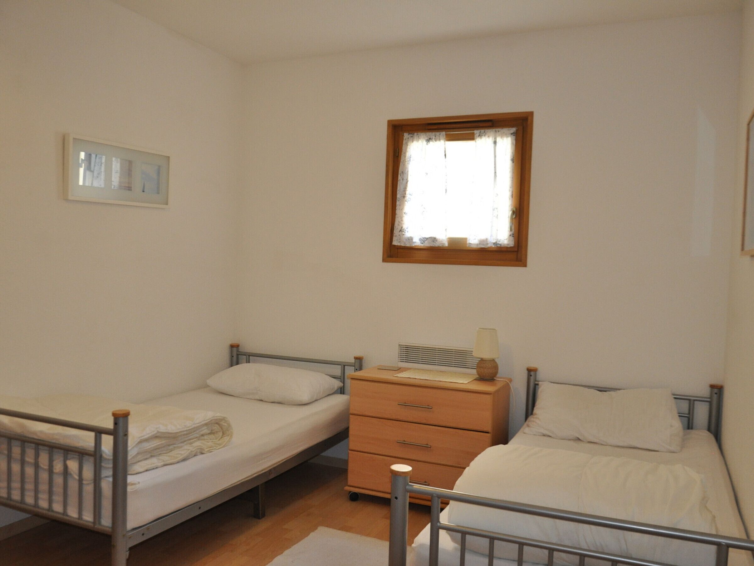 A small, single-room accommodation with two single beds, a wooden dresser, a radiator, and a window with a curtain.