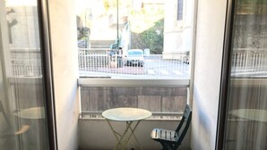 Ein kleiner Balkon mit Tisch und Stuhl, Blick auf eine Straße mit parkenden Autos und ein Gebäude im Hintergrund.