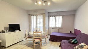 Ein gemütlicher Raum mit Esstisch, Stühlen, einem Bett mit violetter Bettdecke, einem Fernseher auf einem weißen Schrank und einem Fenster mit Vorhängen.
