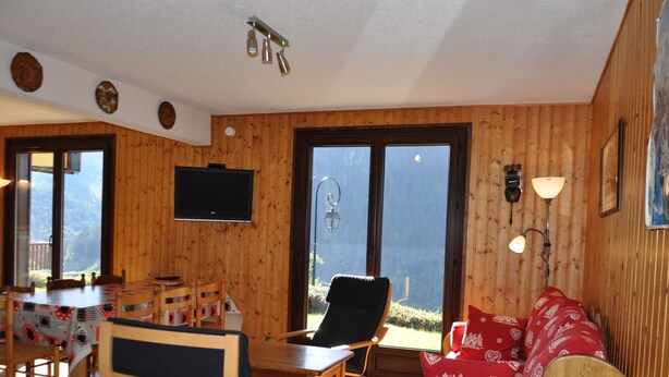 A wooden-paneled room with a dining table, chairs, a sofa with red cushions, a television, and a large window. - Spacious apartment in Châtel with sunny terrace (Châtel)