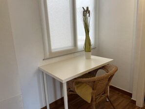 Una piccola scrivania bianca con una sedia in vimini e un vaso con piante essiccate.