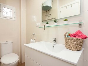 A bathroom with a white toilet, a white sink with a woven basket, a glass shelf with plants, and a mirror above the sink. - Bright 1-bedroom apartment in Málaga center (Málaga)