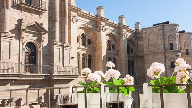 Un edificio de arquitectura clásica, con columnas y ventanas arqueadas. Hay dos copas de vino sobre una mesa, con una valla blanca y flores rosadas al fondo.