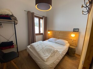 Um quarto com cama, cabeceira de madeira, janela com cortinas, radiador, cabideiro com roupas e um barril de madeira.