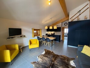 Sala de estar moderna com televisão de tela plana, poltronas amarelas, área de jantar com mesa de madeira e cozinha ao fundo.