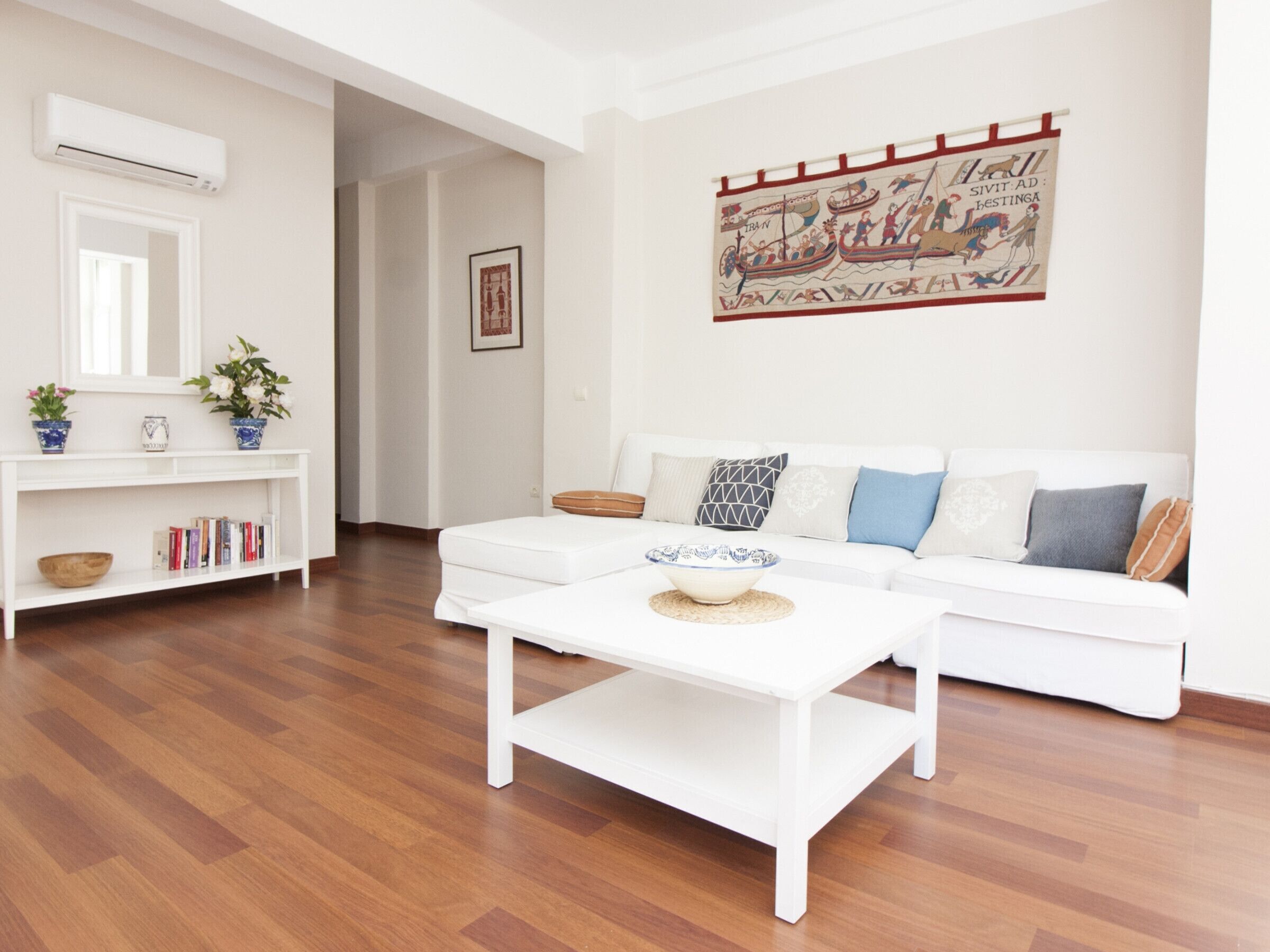 Una sala de estar moderna con un sofá blanco, una mesa de centro y un tapiz colgado en la pared que representa una escena histórica.