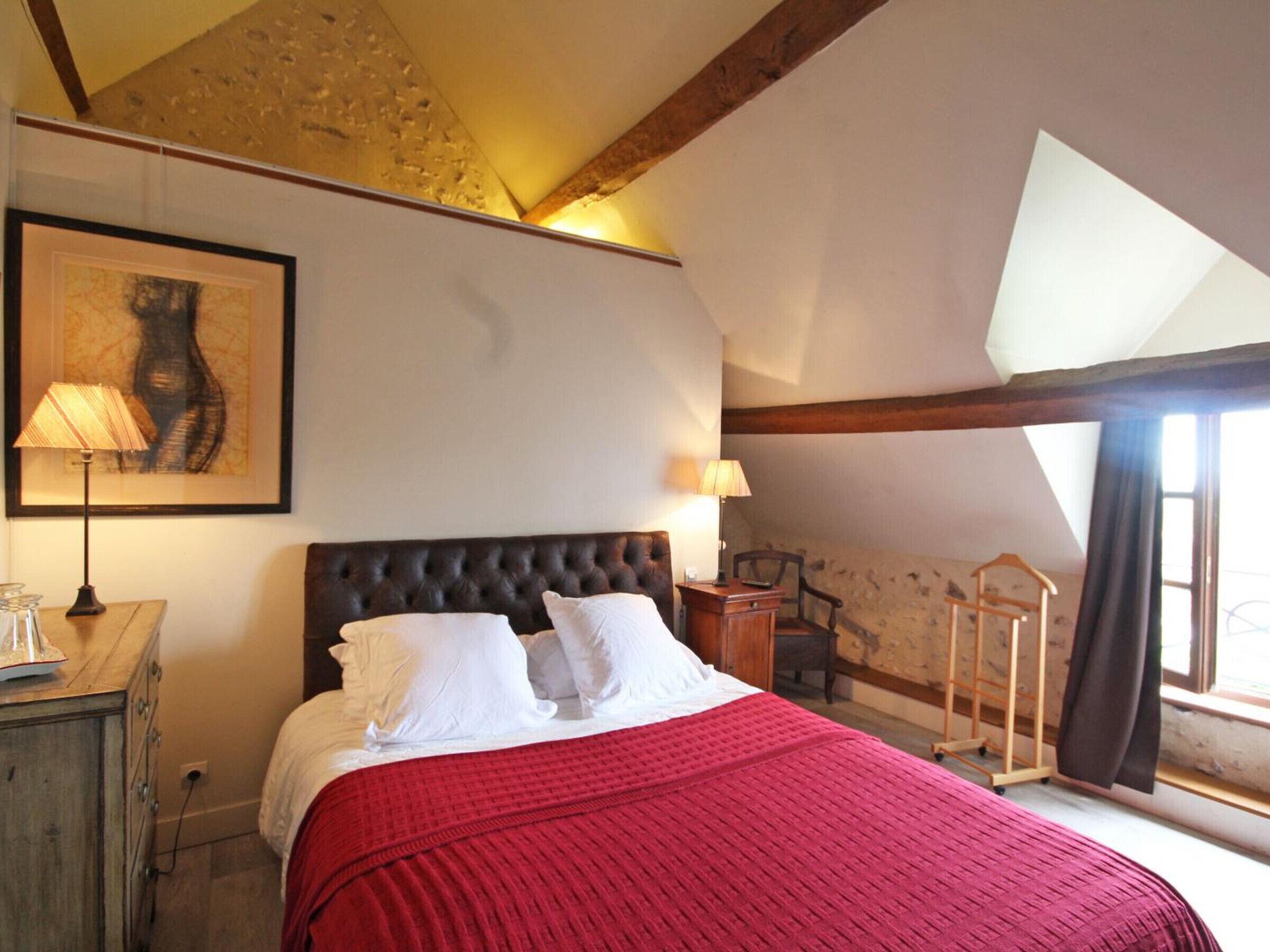 Une chambre avec un grand lit, une tête de lit en bois, une couverture rouge, une œuvre d’art encadrée accrochée au mur et une fenêtre avec des rideaux.