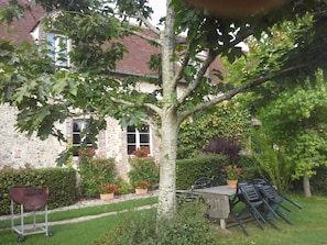 Un edificio de piedra con fachada de ladrillo, un árbol en primer plano y un jardín con plantas en macetas y mobiliario exterior.