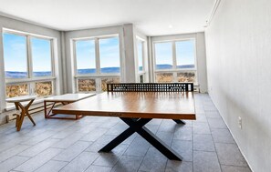 A spacious room with a large table, two chairs, and a bench. There are large windows offering a view of the landscape.