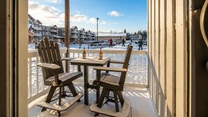 A covered outdoor seating area with Adirondack chairs and a table, overlooking a snowy landscape with buildings and people in the distance.
