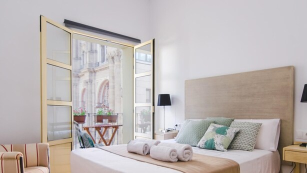 A bedroom with a bed, a chair, a nightstand, a lamp, and a view of a building through a glass door. - Charming Renovated Apartment with Cathedral View in Central Málaga (Málaga)